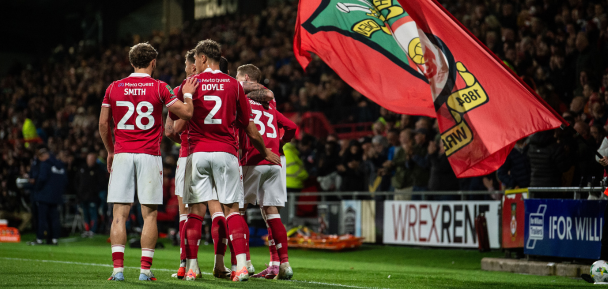 Football players celebrating on the field with a flag, surrounded by fans and advertisements.